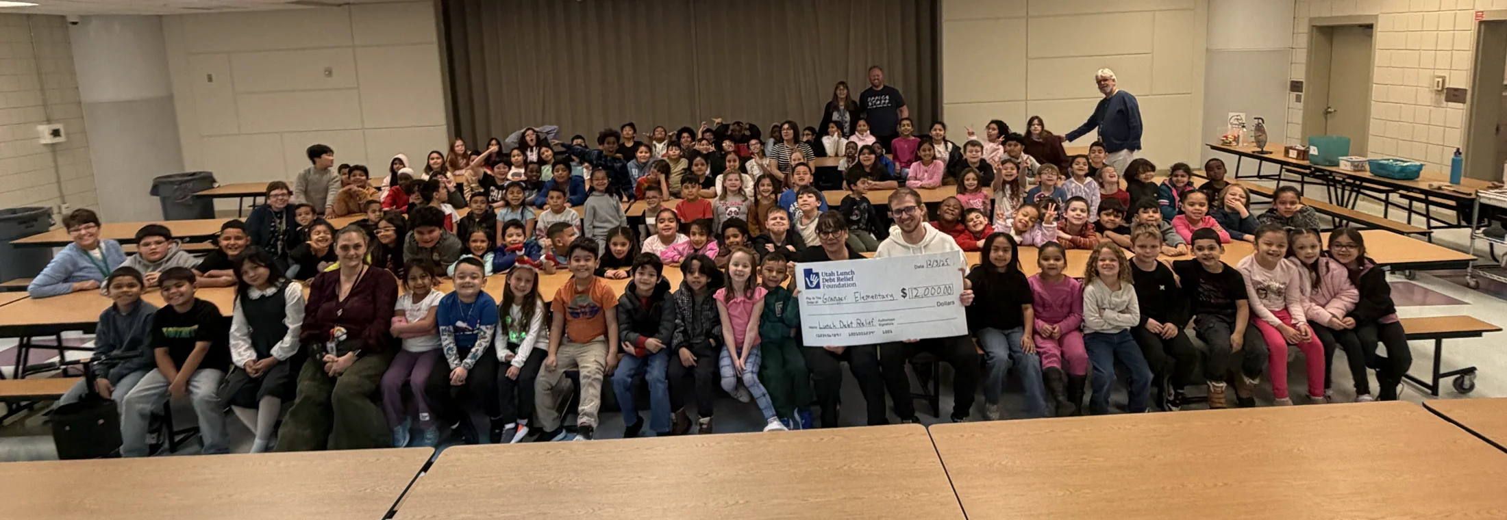 DJ Bracken holding a school lunch debt relief check with students in a lunchroom